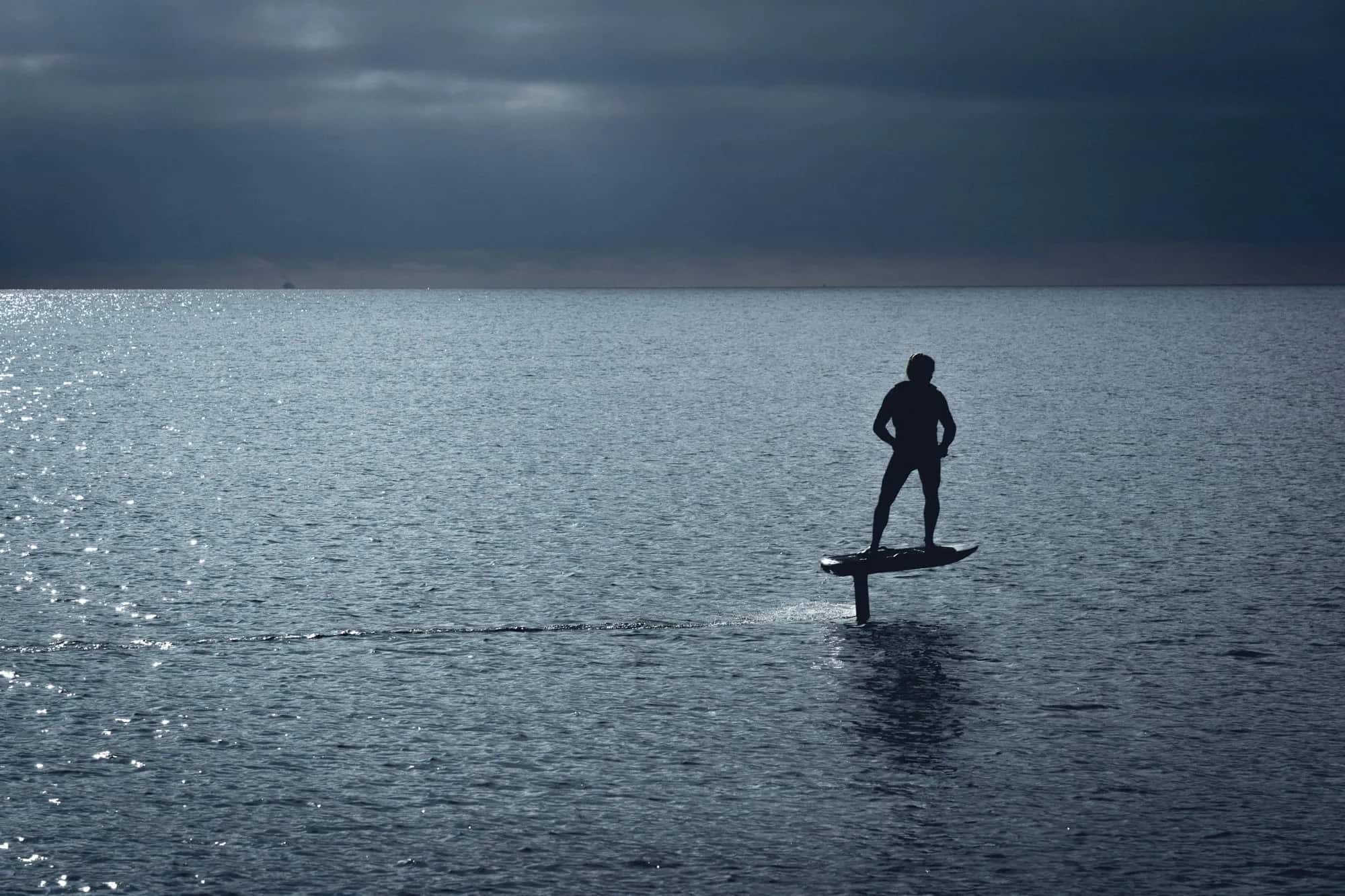Un rider en silhouette, en vol sur un e-foil au-dessus d’une mer d’argent, sous un ciel couvert